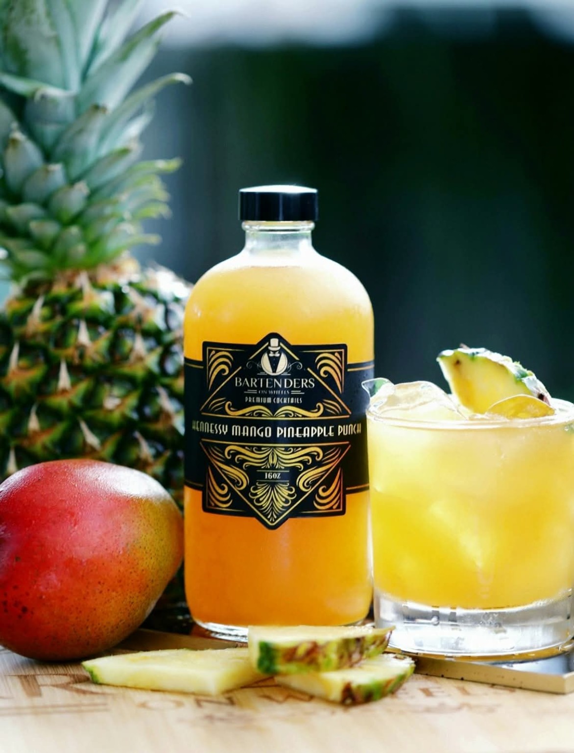Bartenders On Wheels Hennessy Mango Pineapple Punch bottle with fresh fruit and a cocktail over ice.